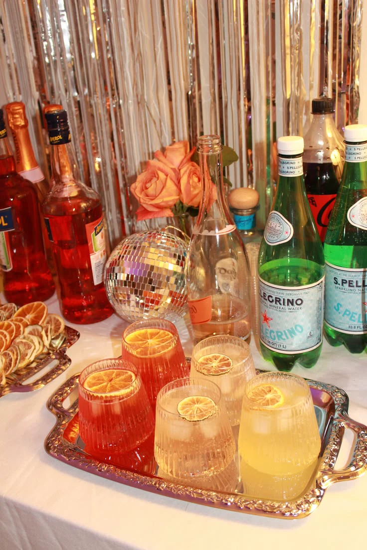 A festive cocktail table with citrus drinks, bottles, flowers and a mirrored disco ball