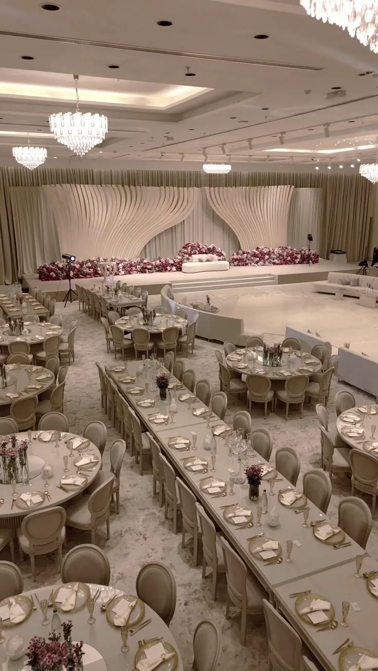 A large ballroom reception with round and long tables, floral centrepieces and a decorated stage